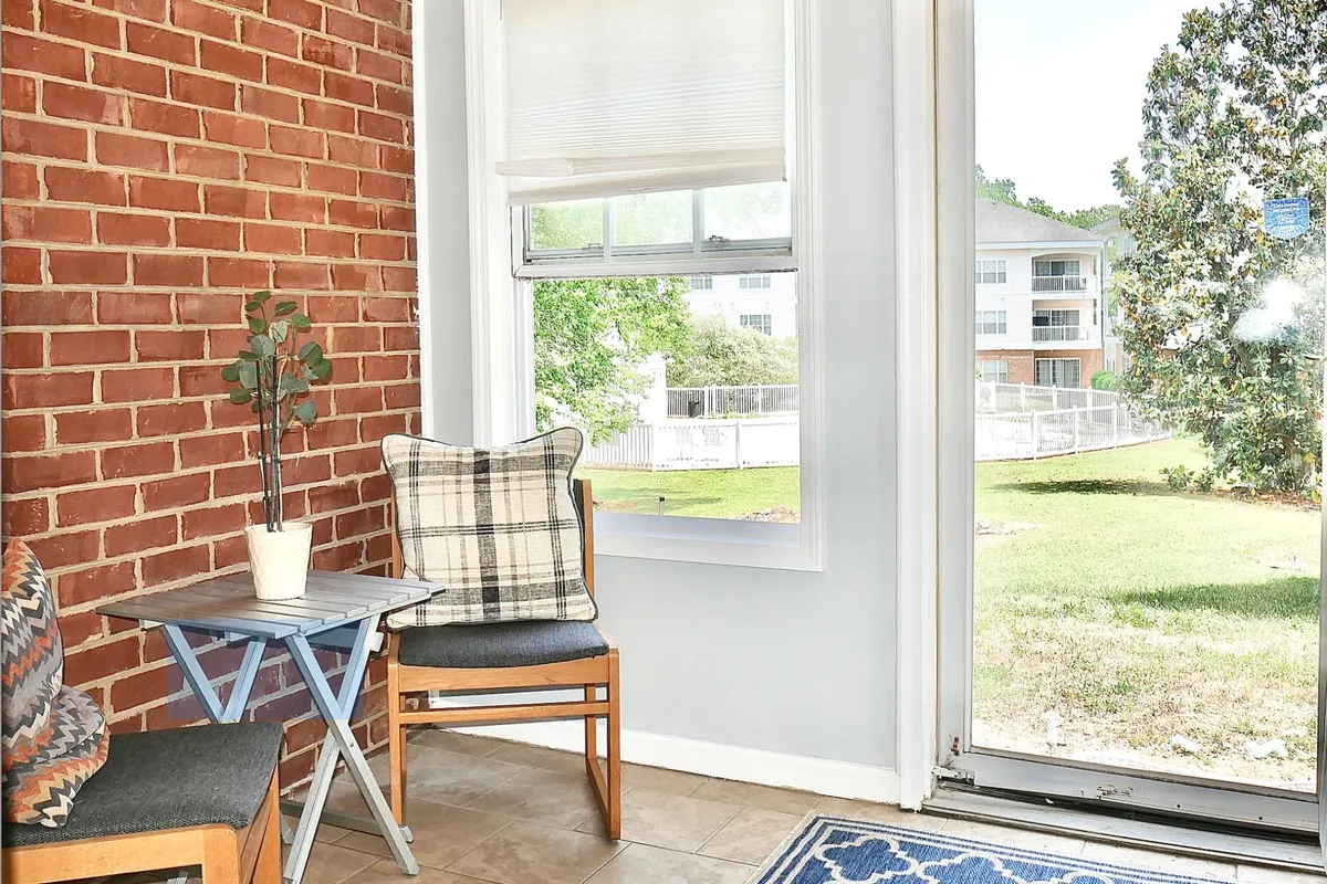 A cozy interior corner with a plaid armchair next to a small table with a plant, set against a brick wall and window. The space is bathed in natural light, ideal for quiet reading, enjoying coffee, or simply unwinding in comfort.