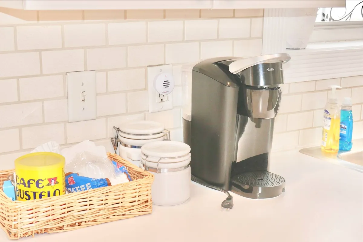 A cozy kitchen corner featuring a coffee maker, a basket of coffee supplies, and white storage jars. The setup invites guests to brew coffee and enjoy a casual morning together.