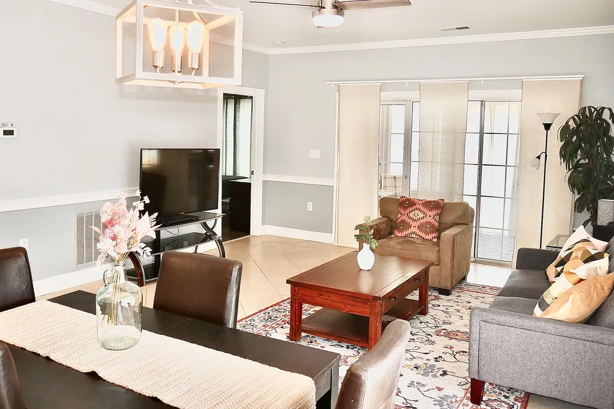 A cozy, open living area featuring a comfortable sofa, a large TV unit, a dining table with a floral centerpiece, and expansive windows with sheer curtains that fill the space with natural light. The arrangement encourages relaxation, conversation, and shared experiences.