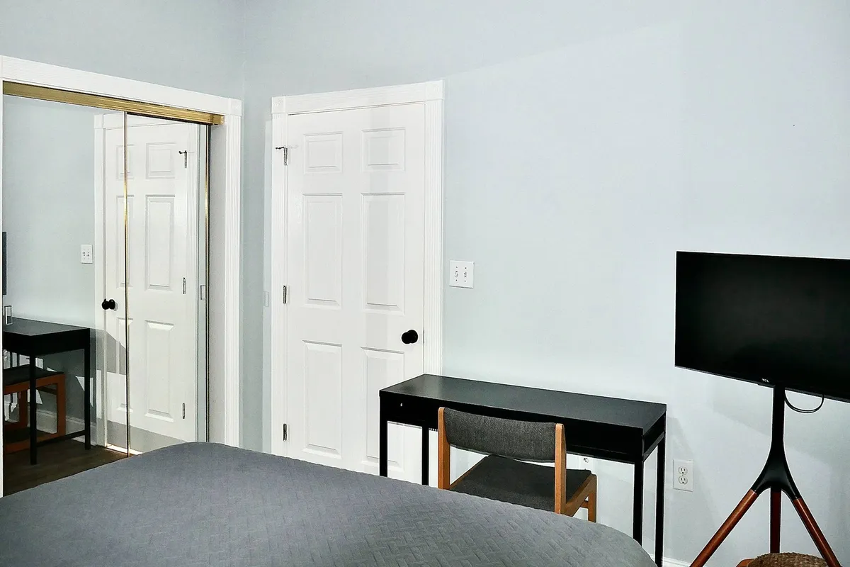 A tranquil bedroom featuring a cozy bed, a dedicated desk and chair set against the wall, a mounted TV, and a large mirror. Soft lighting and neutral decor create an inviting atmosphere ideal for rest, productivity, or downtime.