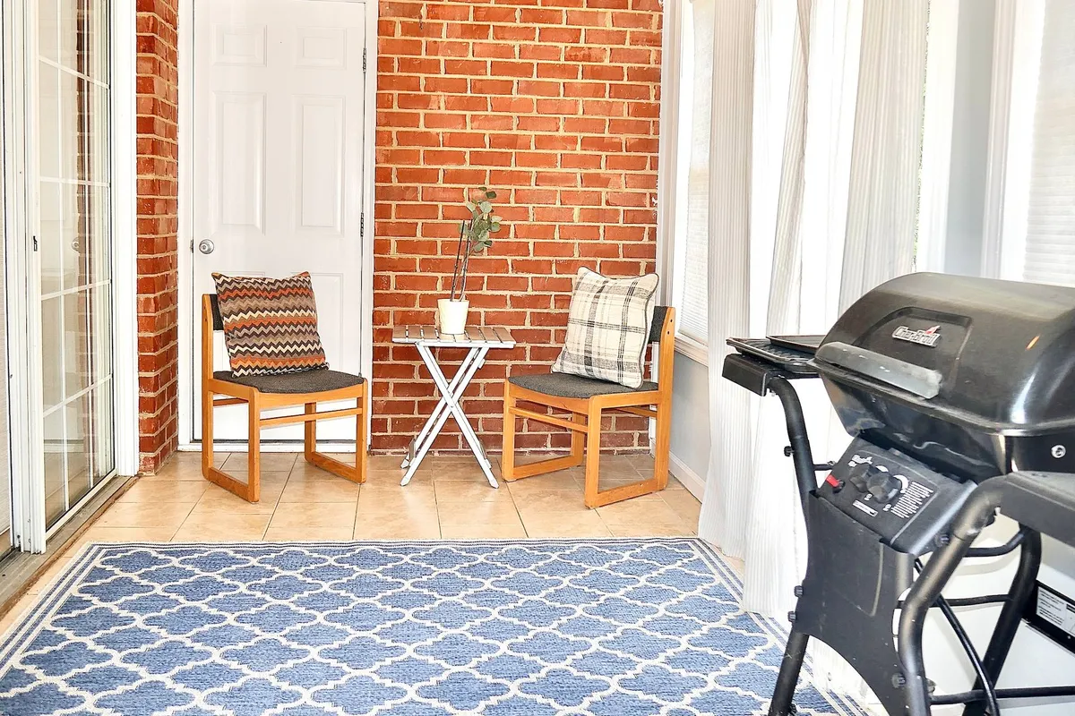 A sunlit sunroom characterized by a charming brick wall, two cushioned chairs, a small white side table, and a blue patterned rug. The setting exudes coziness and is perfect for enjoying a quiet moment or socializing in a sunny, relaxed atmosphere.