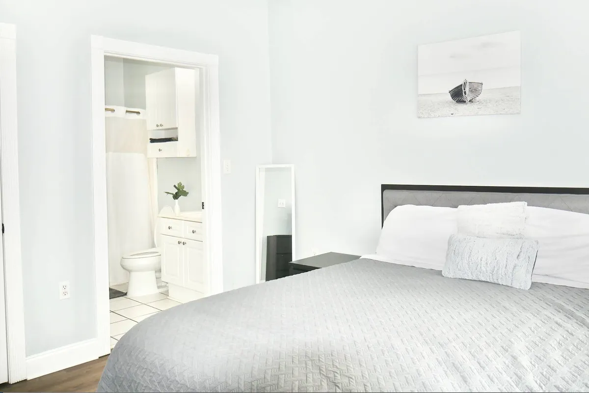 A serene bedroom with a textured bedspread, soft lighting, and a piece of framed art depicting a boat above the bed. Through a partially open door, a glimpse of the bathroom area is visible, connecting sleeping and bathing spaces in a cohesive suite.