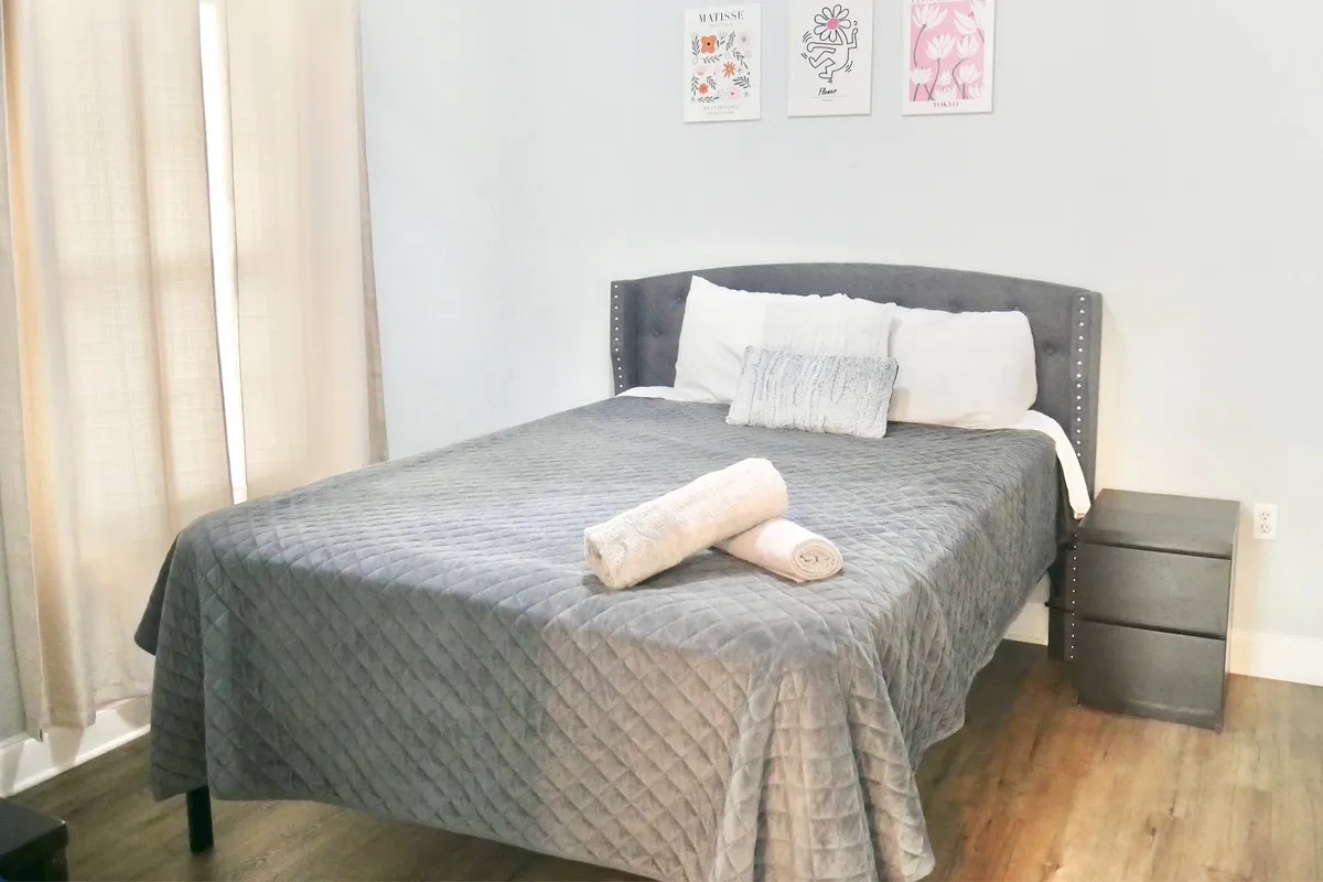 A cozy bedroom with a neatly made bed, decorative pillows, plush towels on the bed, and tasteful wall art. Sunlight streams in, creating a calm and inviting atmosphere, ideal for restful mornings or evenings.
