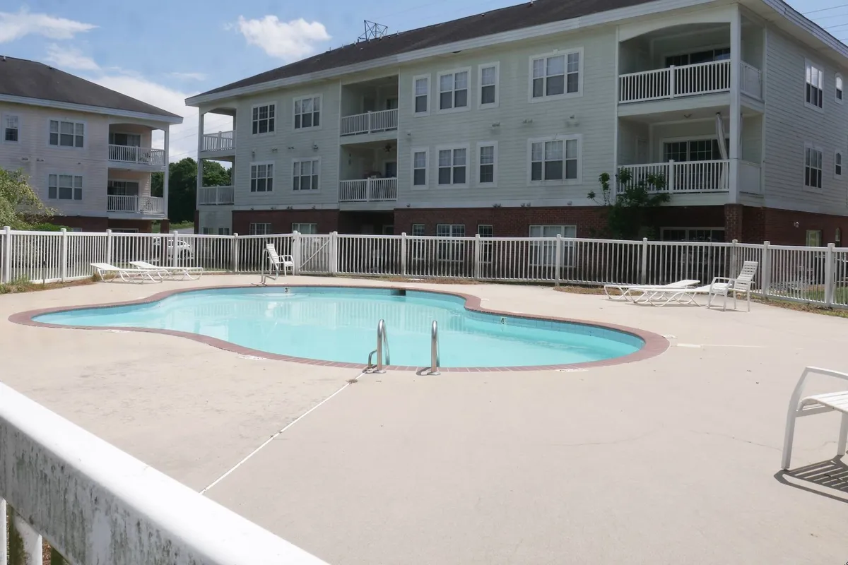 A vibrant exterior shot of the community's swimming pool, surrounded by apartment buildings and plenty of poolside lounge chairs. The setting is bright, inviting, and clearly designed for sunbathing, social gatherings, and leisurely swims. The image captures the heart of the resort-style atmosphere and outdoor amenities.